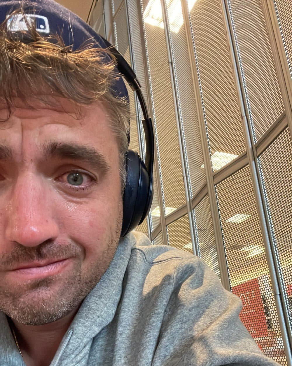 Adam, in a grey hoodie and headphones, eyes red, half-smiling at the camera in an airport.